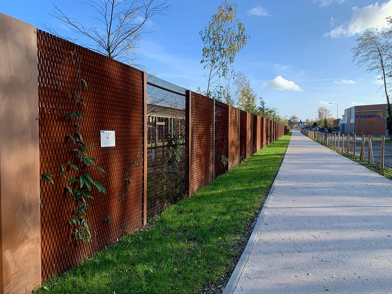 Allée verte bordée d'une clôture Corten près d'un E.Leclerc Long chemin piétonnier longeant une clôture en métal rouillé (Corten) avec une vigne grimpante et un panneau blanc. Un bâtiment E.Leclerc DRIVE est visible au loin sous un ciel bleu clair.